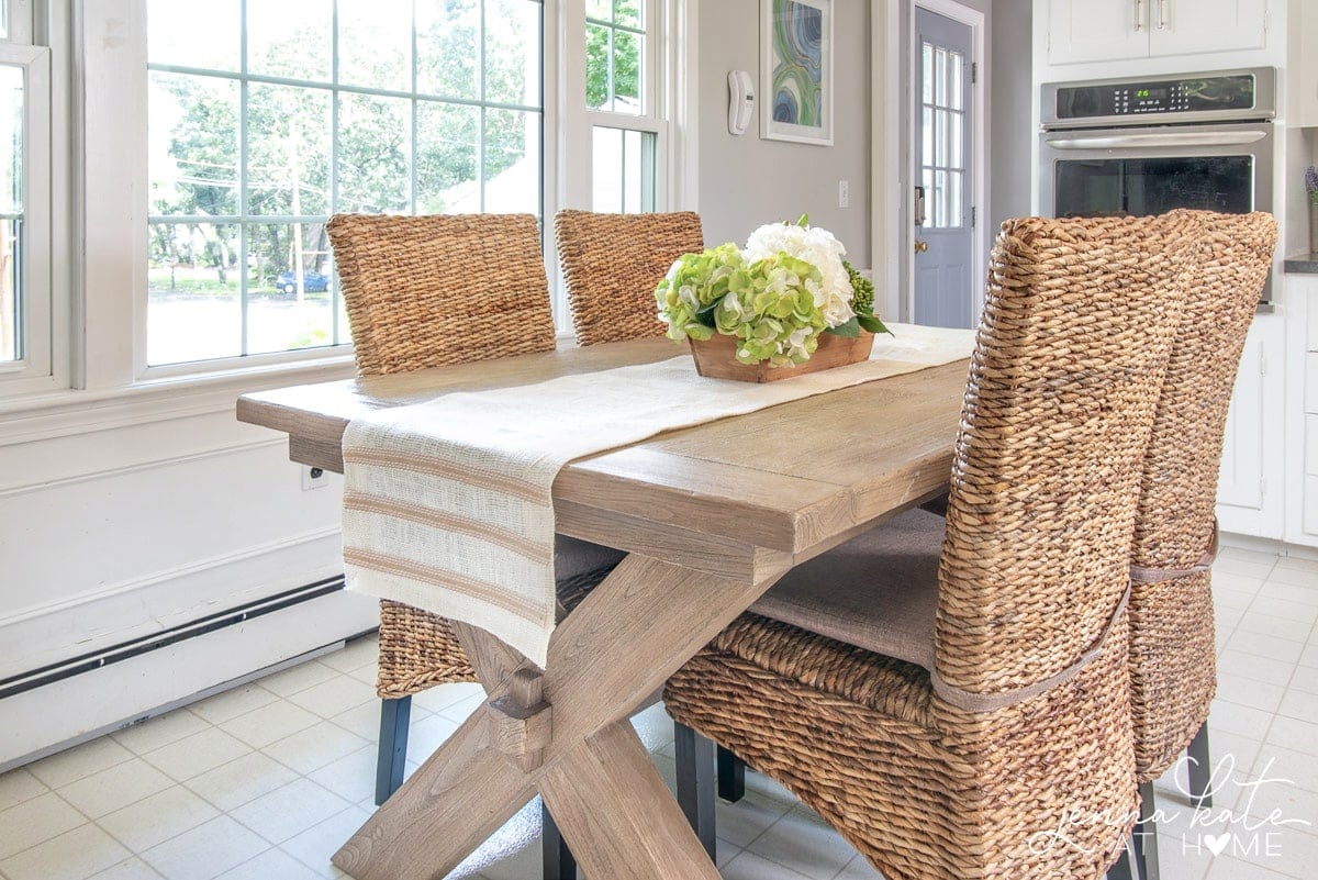A light brown, wooden dining table with wicker high-back chairs and a beige/white table runner