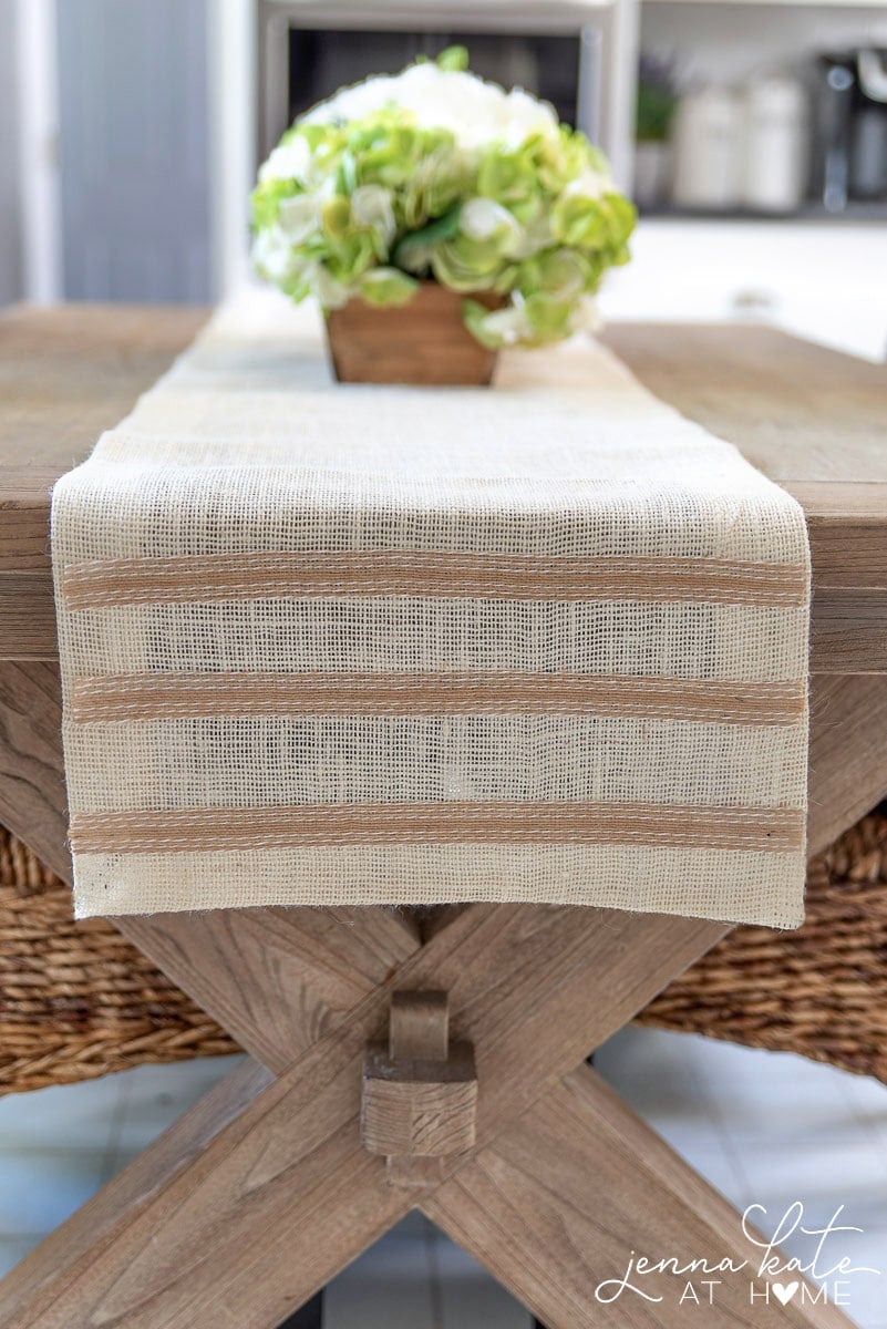 The finished burlap runner, now placed on top of a wooden table in the kitchen, with a low green centerpiece in a wooden plant holder