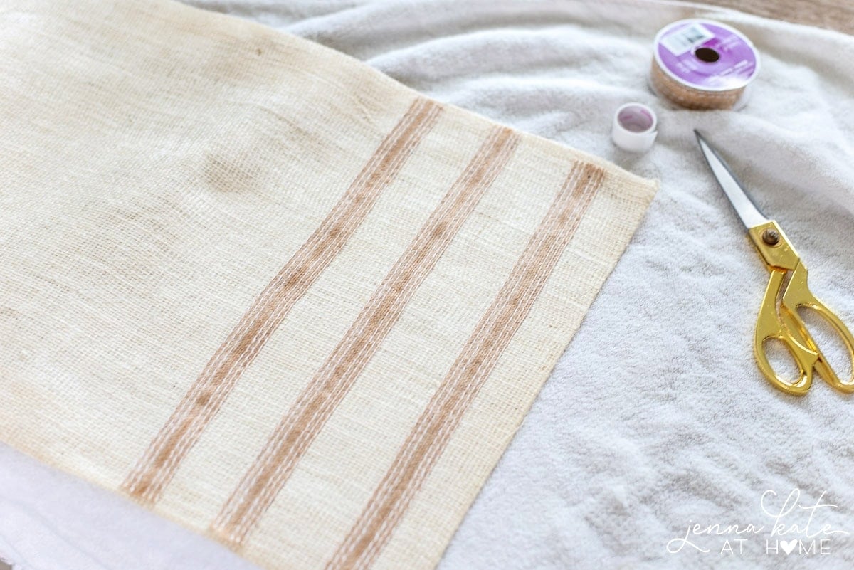 The burlap runner resting on a towel, near the fusing tape and scissors, after the decorative burlap ribbon has been ironed on 