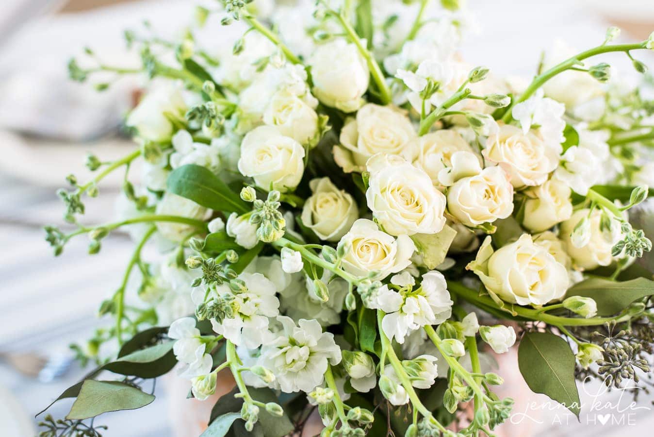 A close up of a large arrangement of white flowers and greenery in a vase on a table