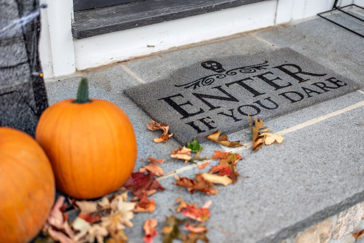 A grey, outdoor welcome mat with the words \"Enter if you dare\" with 2 orange pumpkins and a scattering of fall leaves nearby