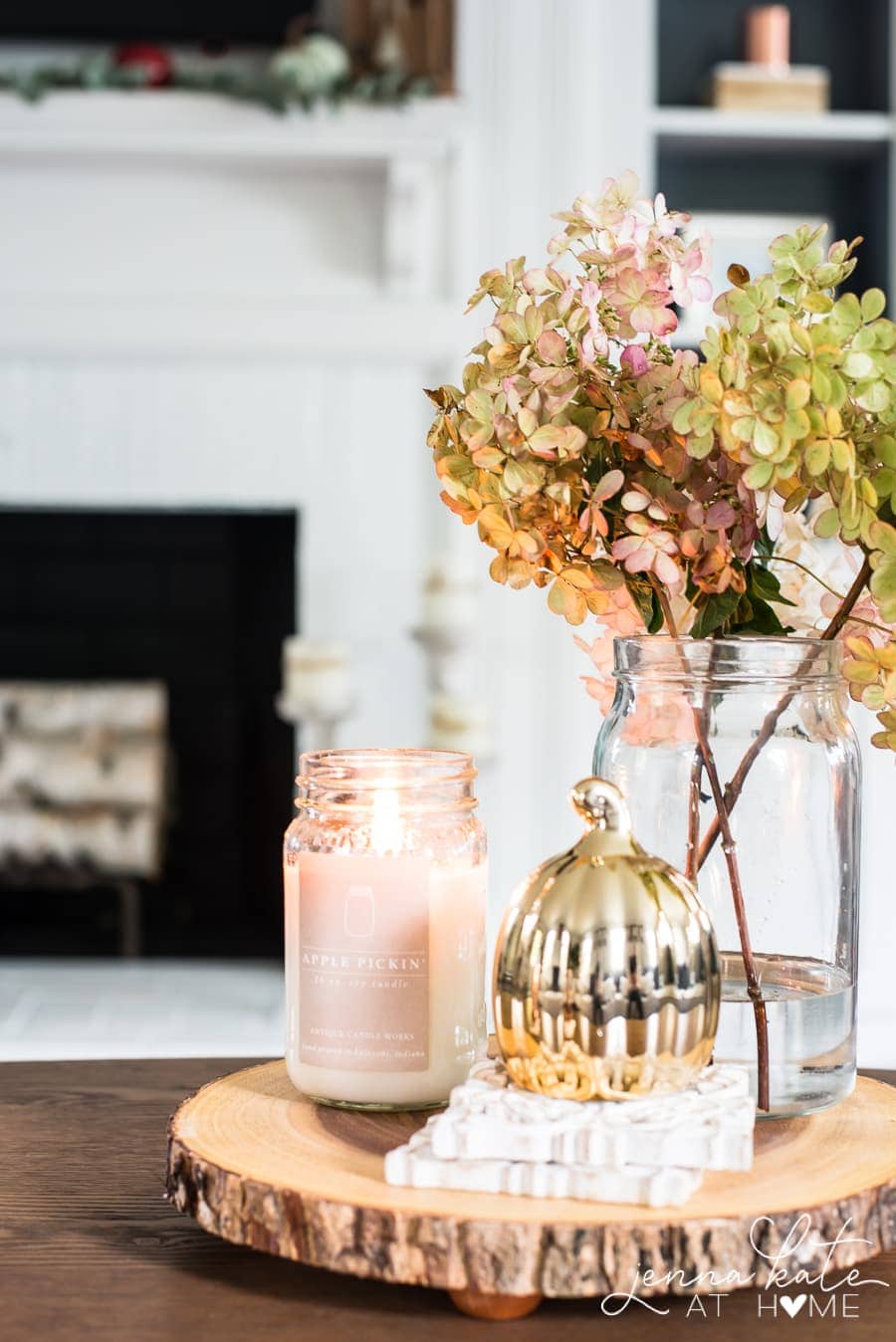 A rustic wooden tray holding a clear vase of fall greenery, a pink candle and a golden pumpkin resting on white, textured coasters