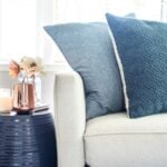 The corner of a light grey couch, with two dark blue pillows, near a metallic blue side table and a copper vase of flowers