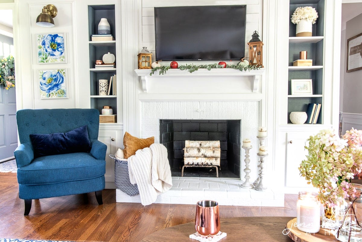 A fireplace mantel with lanterns on top, neutral decor on either side and a tall, blue armchair on the left