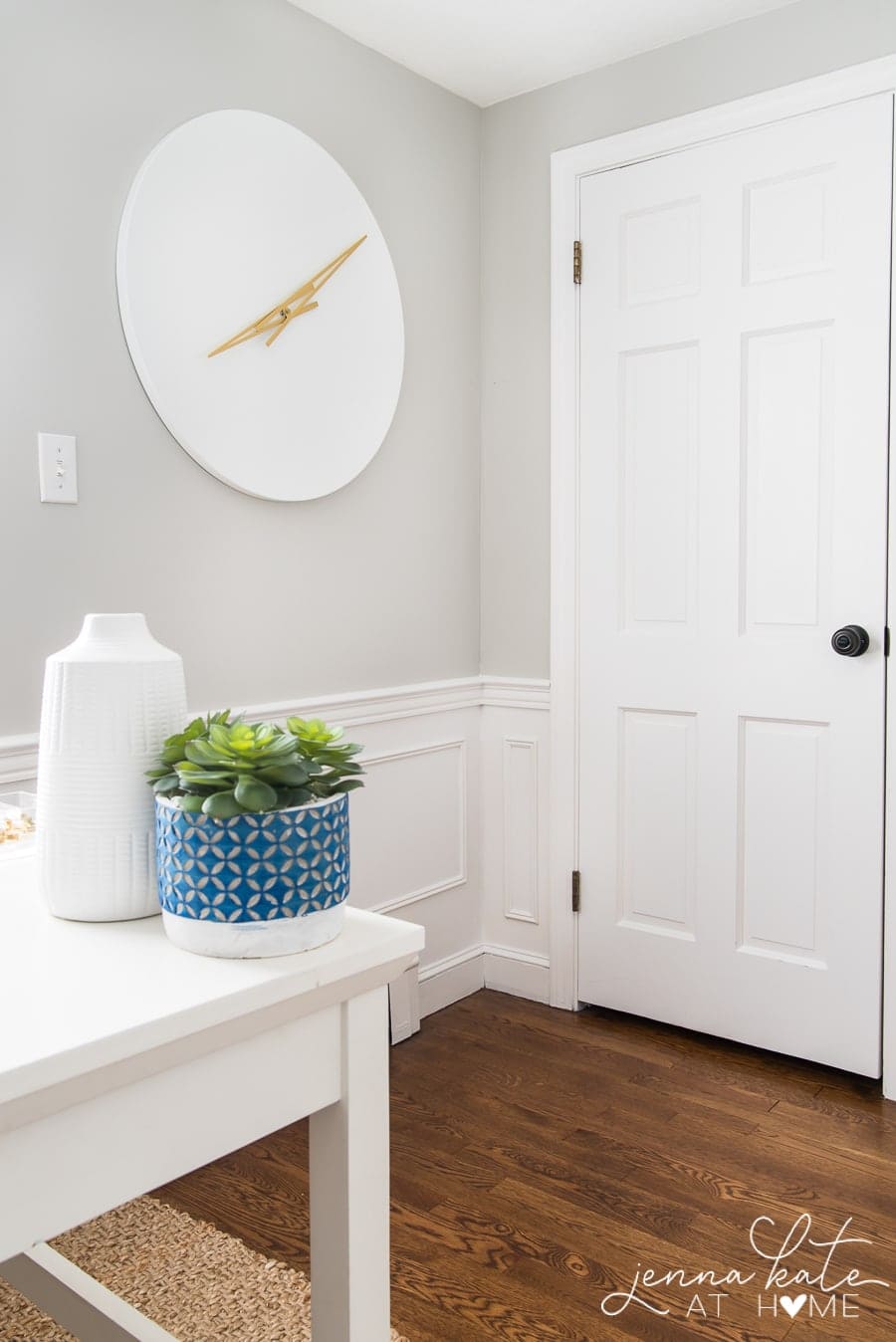 The edge of a desk, holding a potted plant and tall, white jug, with a large, white clock with gold handles nearby, and the door in the background