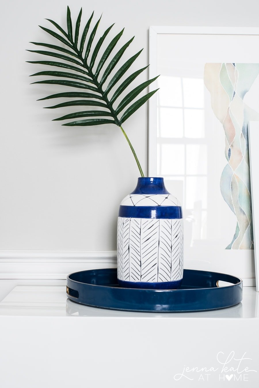 A white jug with painted dark blue line patterns, resting on a circular blue tray, holding a single palm frond near large framed art