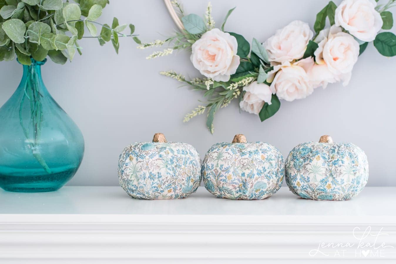 Three pumpkins of the same size, wrapped in vintage patterned paper, near a clear blue vase of greenery and in front of a light pink floral wreath 