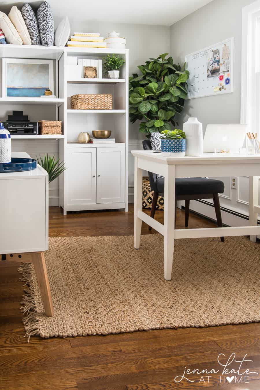 A room with mostly white, wooden furniture (desk, side table and shelf in rear), a black desk chair and brown rug on wood flooring, with a large plant in the corner