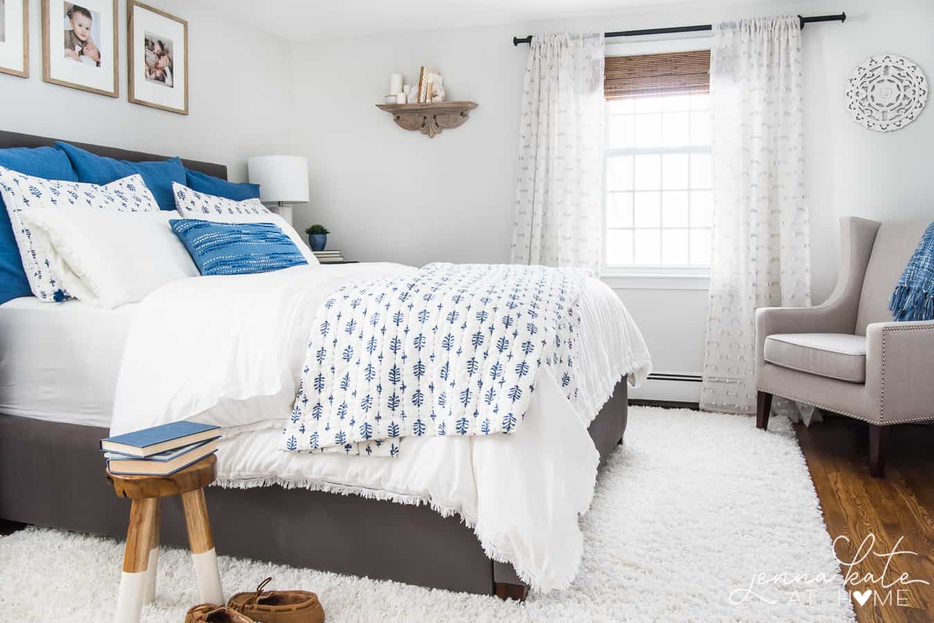 A bedroom with plush, white area rug, a bed with blue and white bedding, and a large window in the background with sheer, white curtains