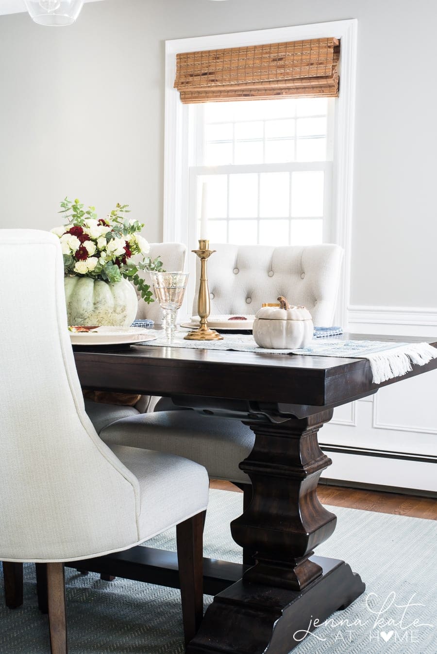 A hefty wooden dining table, dark brown, with off-white, upholstered chairs, and a light green pumpkin centerpiece.