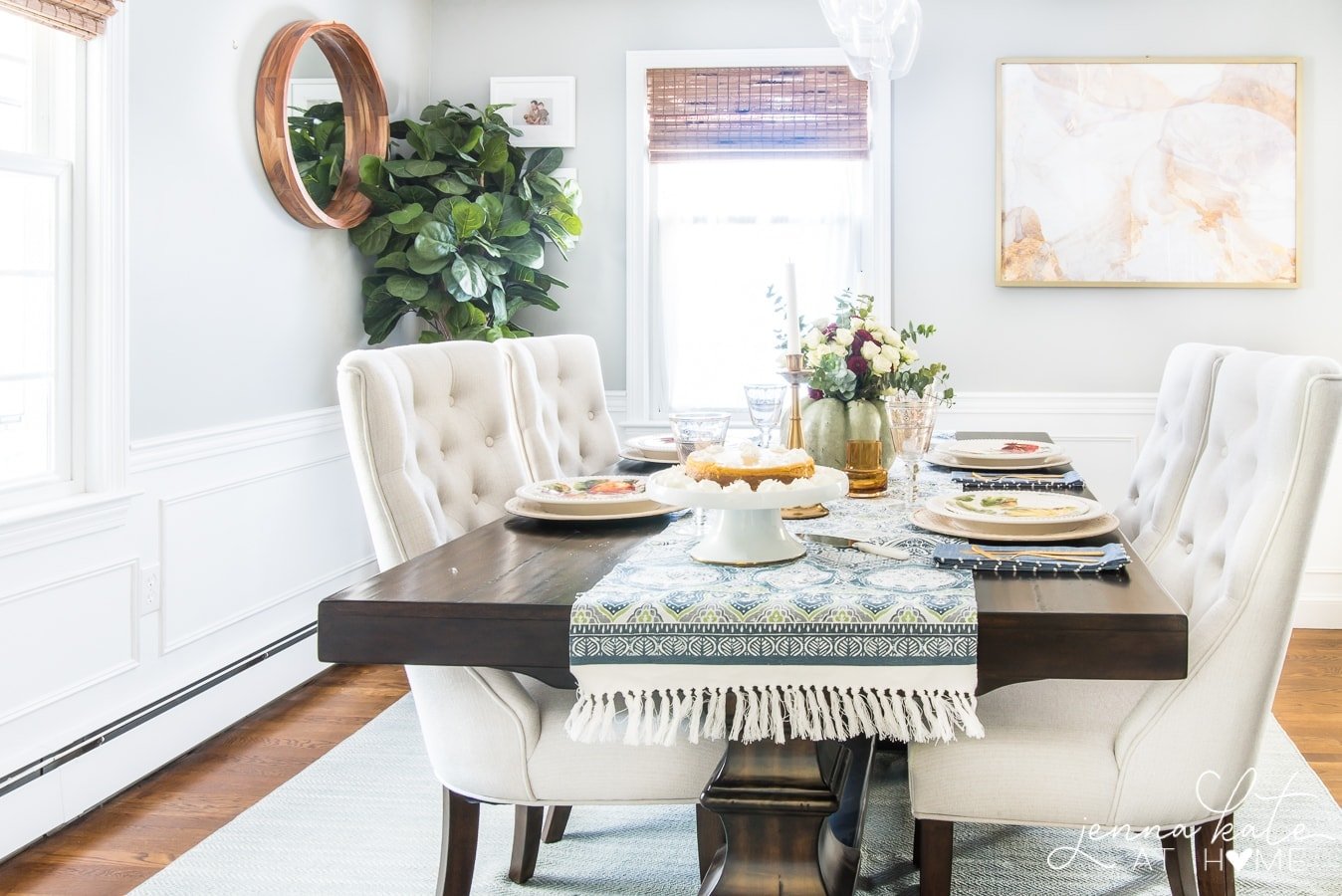 A side view of the dining table with a blue/white runner, pedestal tray with a cake, fall table settings and a light green pumpkin holding flowers.