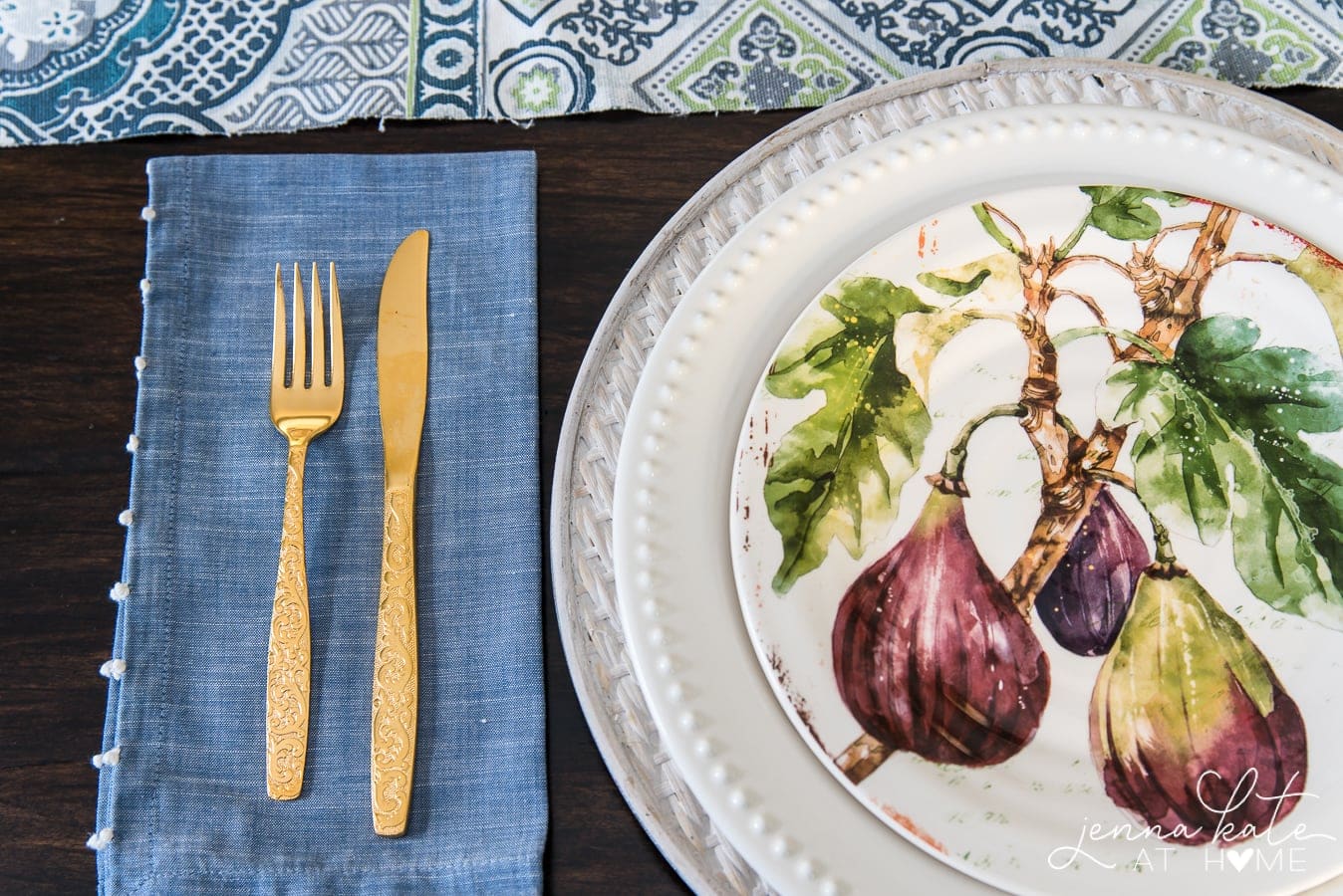 A close-up, top view of the place settings with 1 decorated plate (diagram of figs), one plain white plate and a charger, near a blue napkin with gold utensils