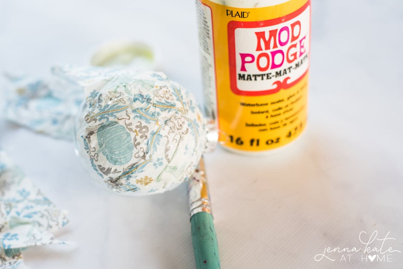 A bottle of Mod Podge, a paint brush and the ornament covered with a blue and white paper napkin