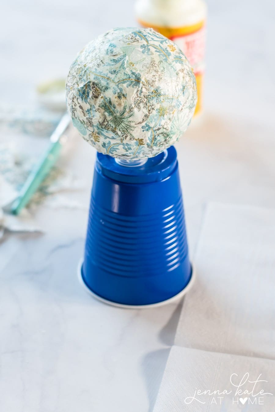 Resting the ornament on a blue plastic cup to let it dry; a paint brush and Mod Podge bottle in the background