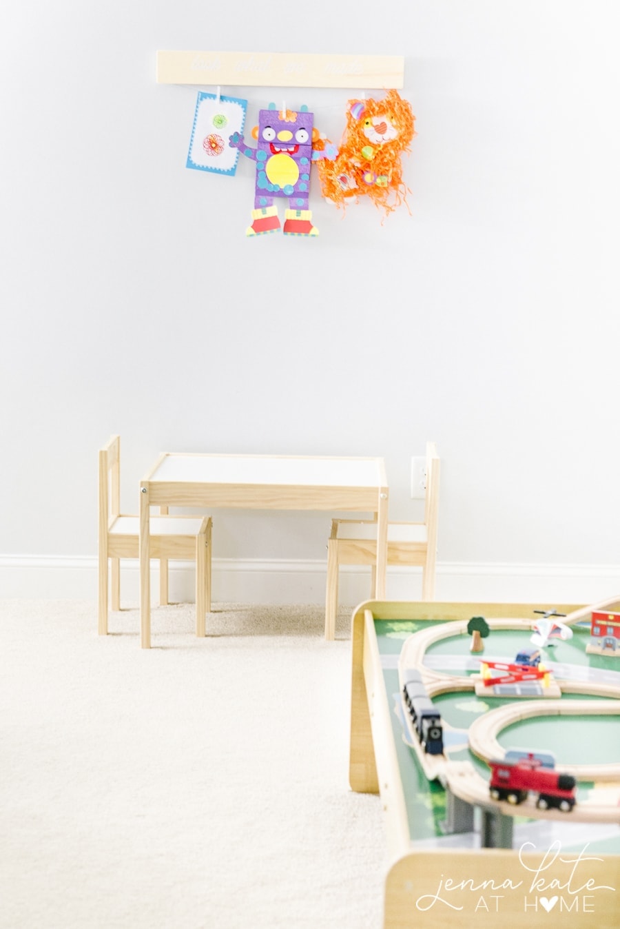 A child\'s playroom with a small table and two chairs, a wooden train table and various artwork hanging on a wooden display