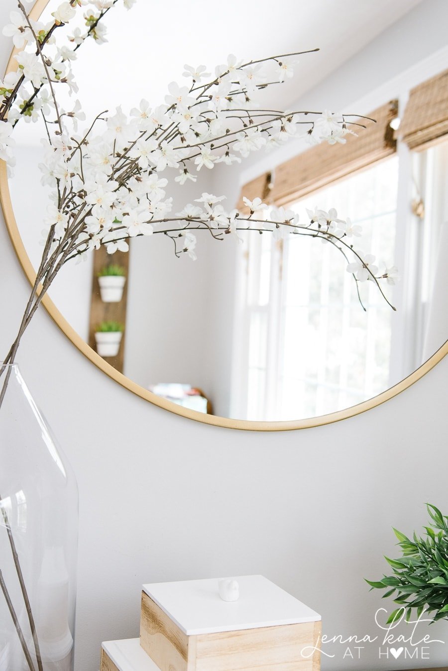 A close-up of a large, round mirror reflecting a trio of plant pots and wooden window blinds in the room