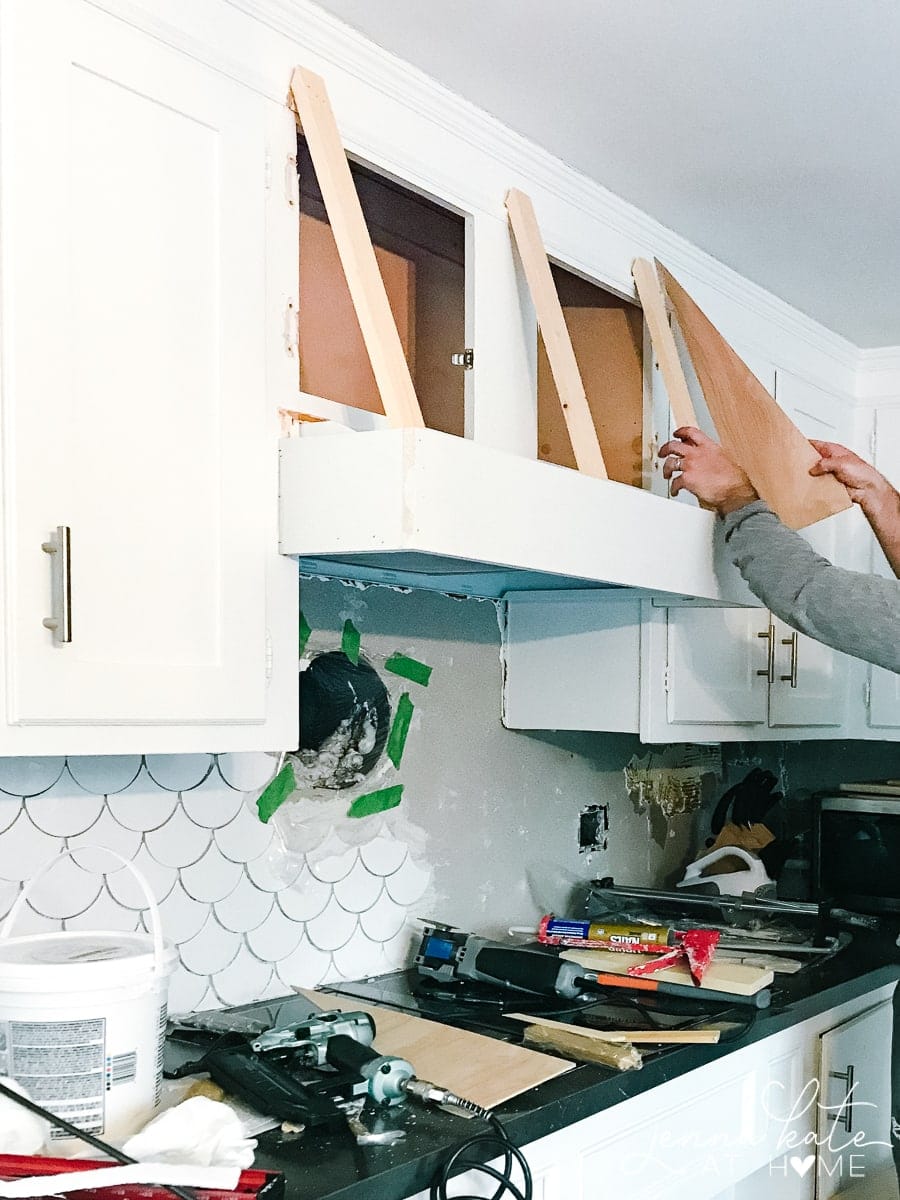 A person holding an triangular piece of wood against the upper cabinet to figure out how the hood cover will be angled