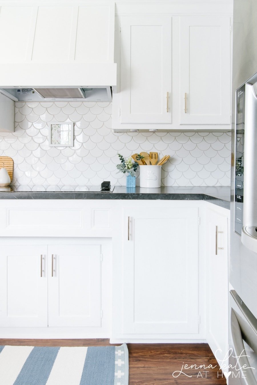 A stovetop built into the cupboard with decorative hood, with stainless steel fridge nearby, white scalloped backsplash with white cabinets