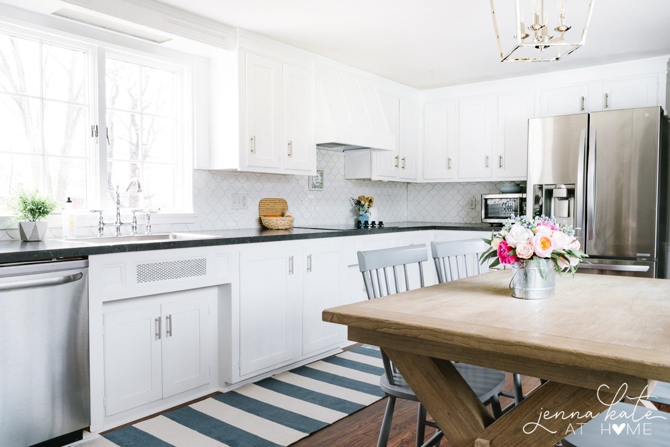 A kitchen with white cabinets, silver hardware, a rustic wooden table and grey chairs, and a blue and white rug on wooden floors