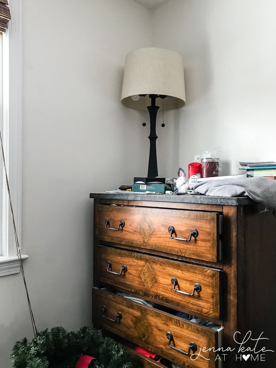 A tall, brown dresser with a beige & black lamp and a clutter of items on top