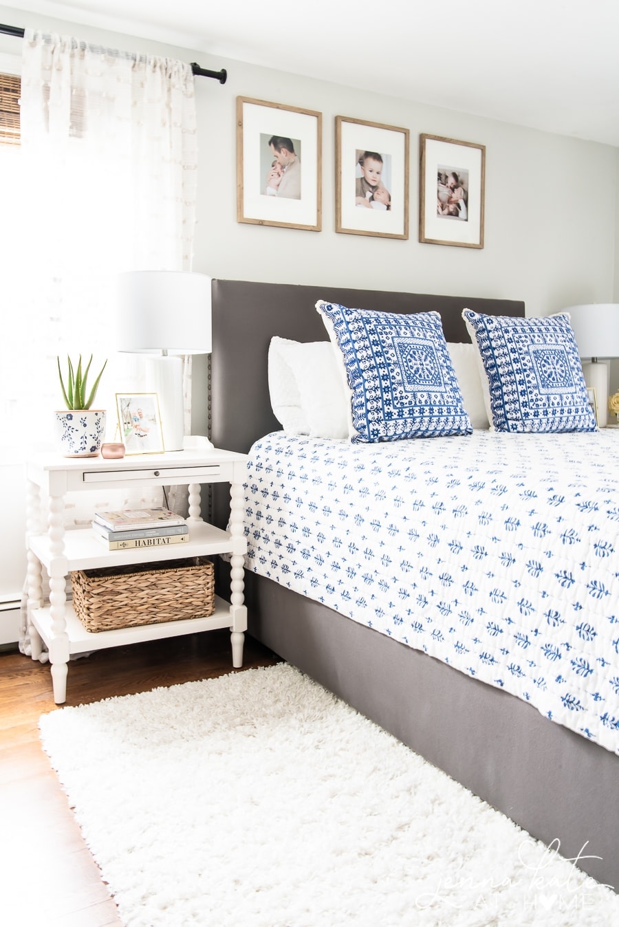 A bed with a grey headboard, blue & pillows and bedding, a white nightstand and white rug.