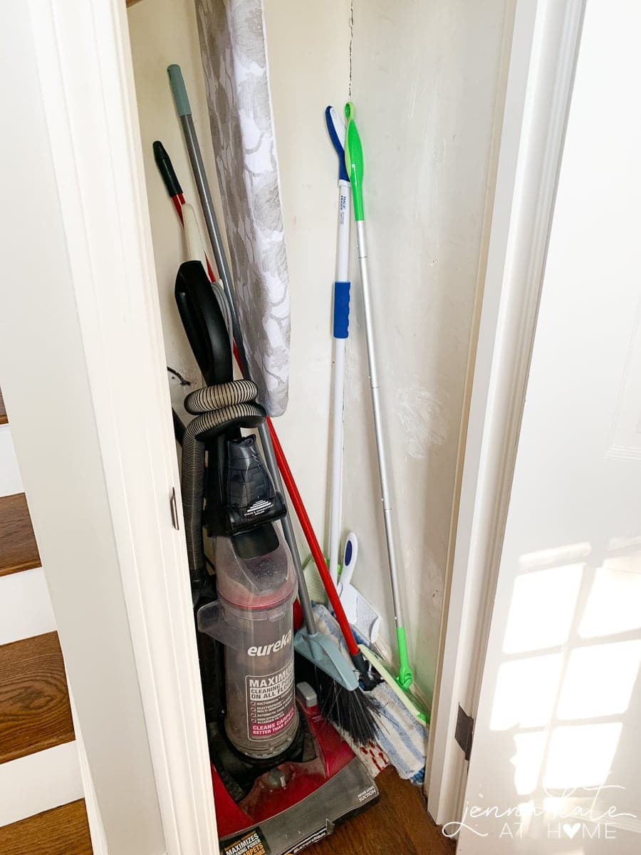 A broom closet crowded with a vacuum, ironing board and various mops and brooms.