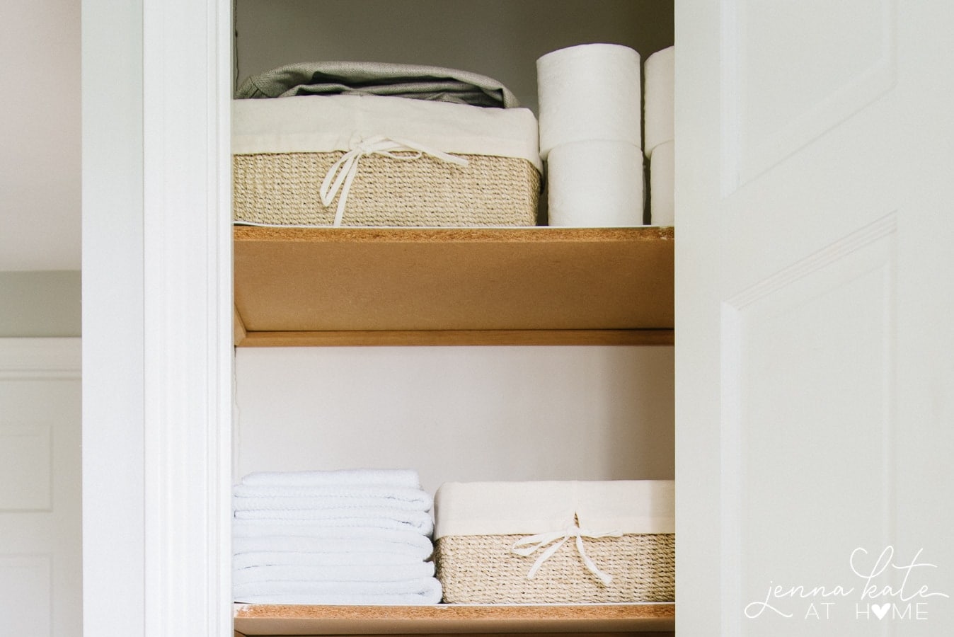 Two closet shelves with beige baskets, stacks of hand towels and rolls of toilet paper
