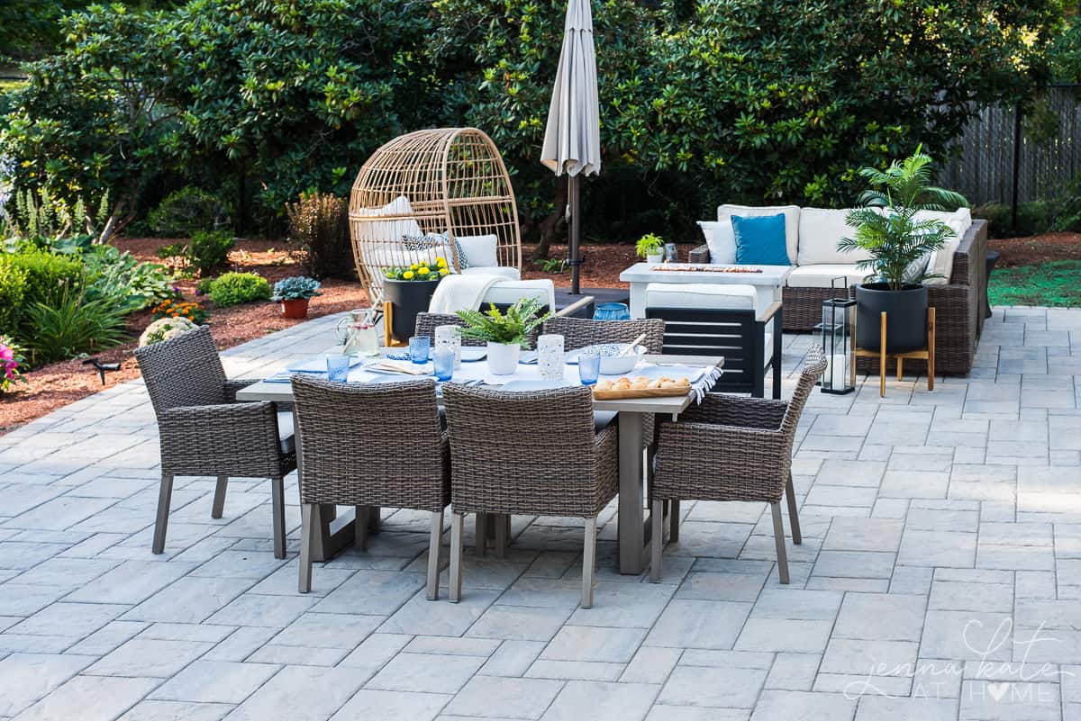 A grey stone patio area with an outdoor table and wicker chairs, with blue and white place settings and other outdoor seating nearby