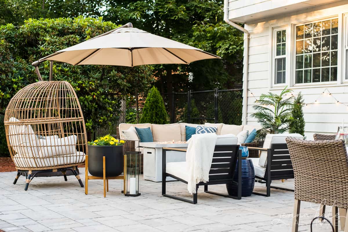 A grey stone patio with sofa set, beige umbrella, and single chairs nearby with white cushions