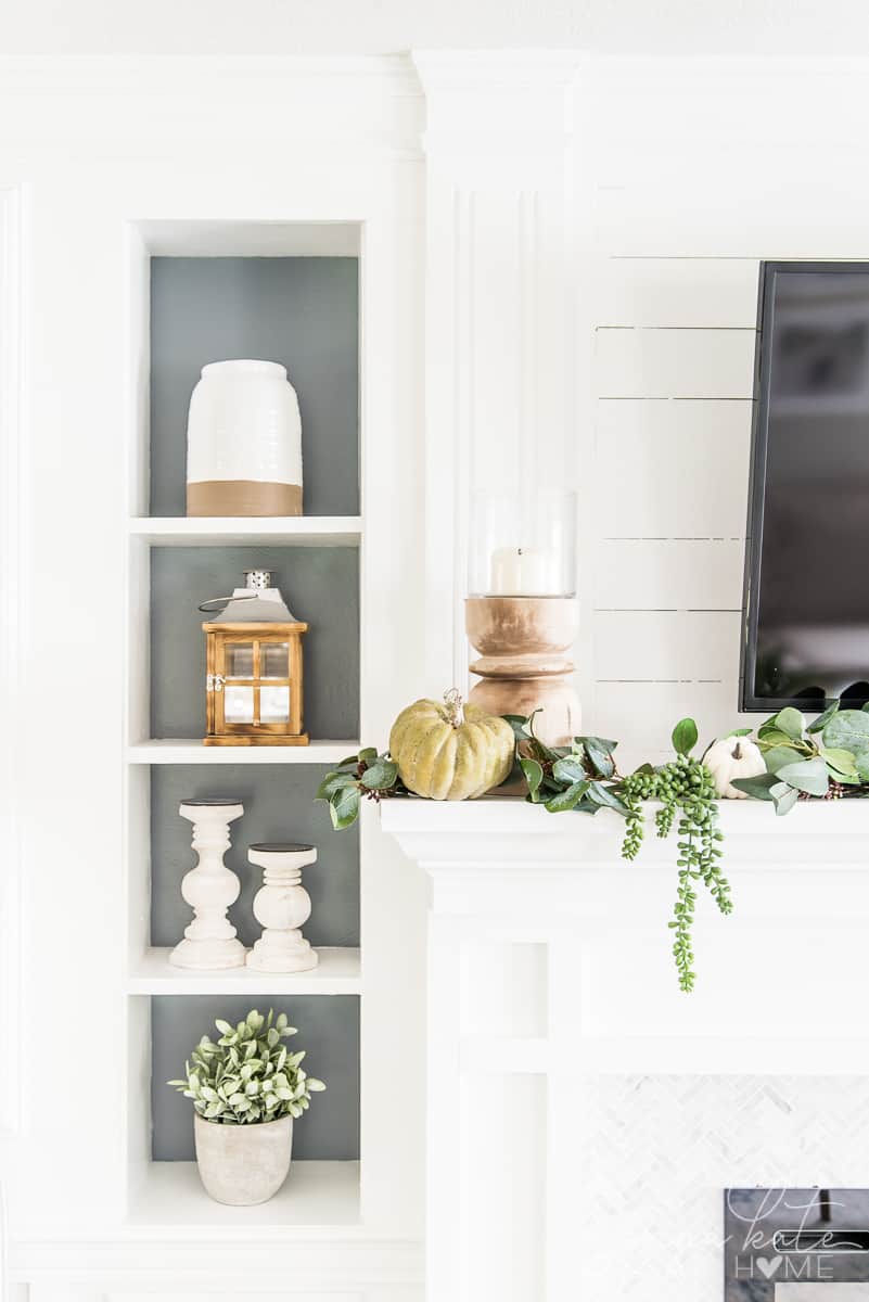 Close up shot of the side of a mantel with a candle stick, faux pumpkin, and greenery draped down the side. A bookcase to the left with various neutral decor elements.