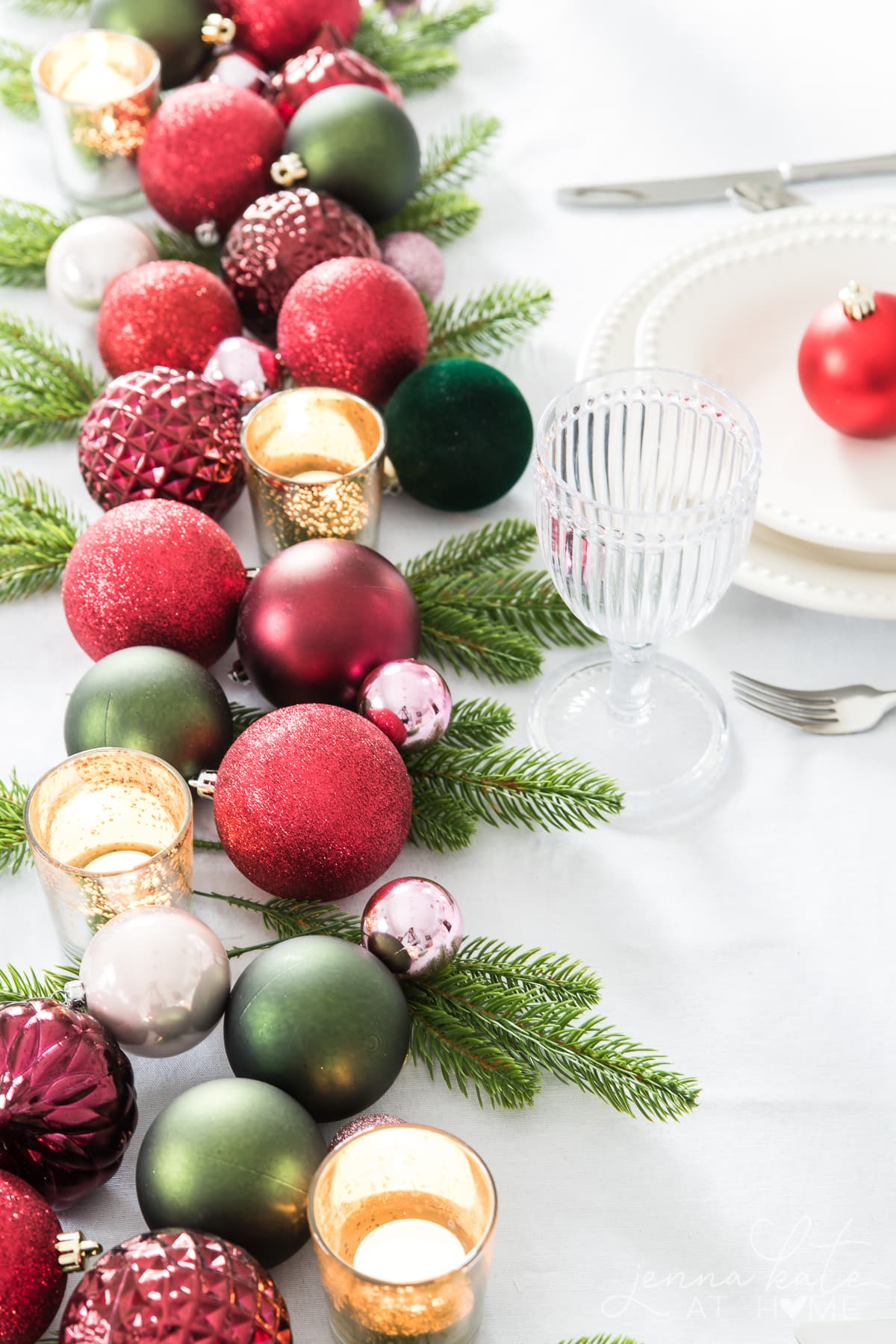 runner of Christmas ornaments, votive candles, and faux greenery on a table