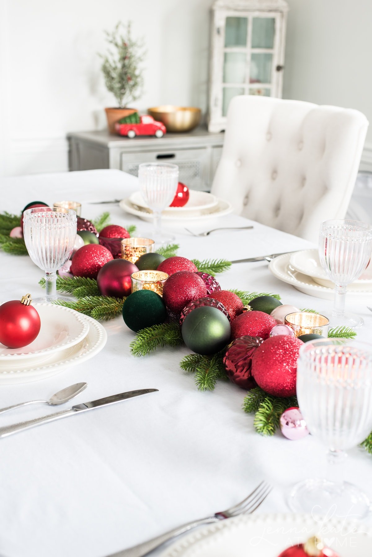 red and green Christmas ball ornaments on a table with gold votive candle holders and artificial greenery