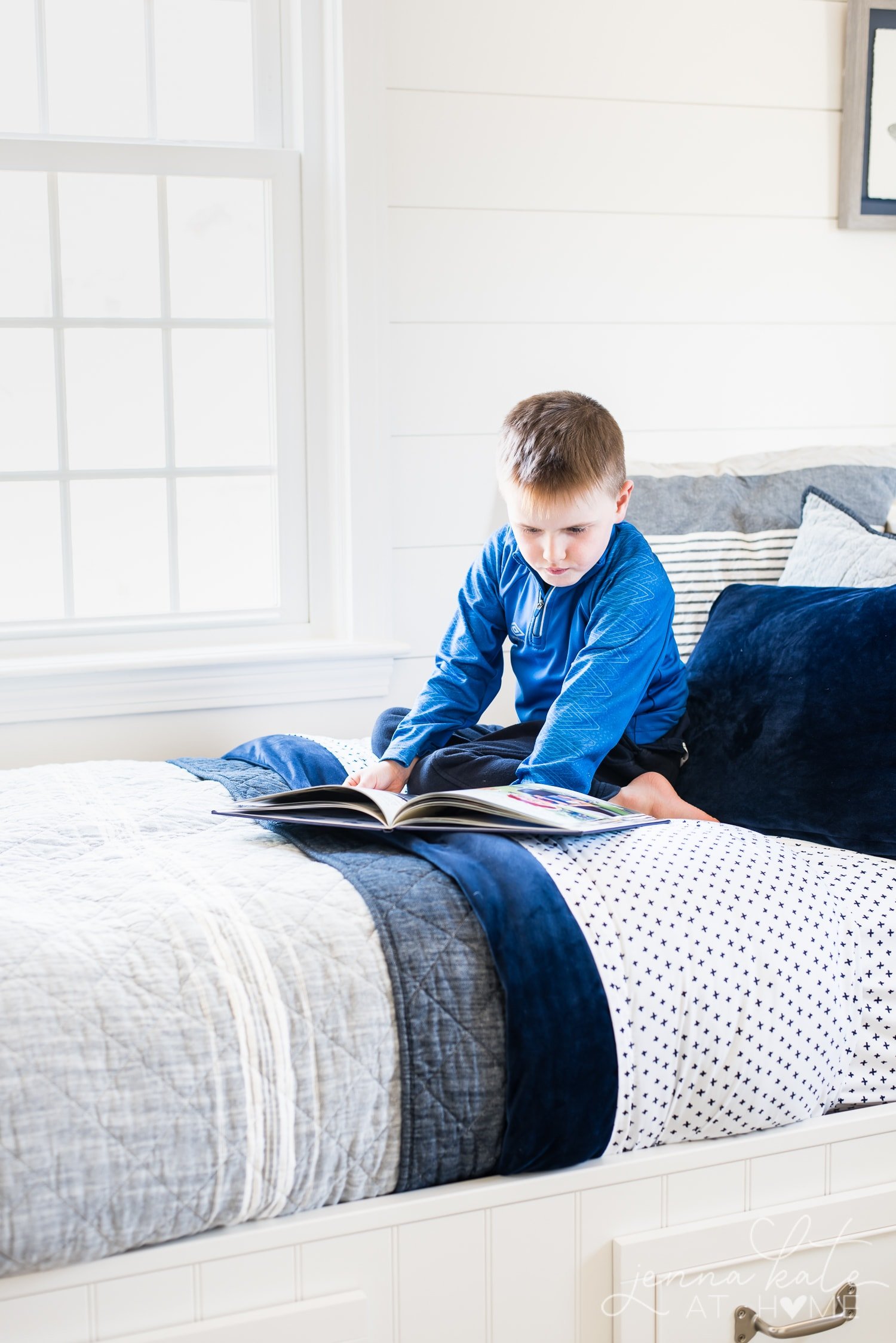 Young boy reading on the bed with layered bedding - small print bedsheets, solid navy duvet cover and gray quilt.