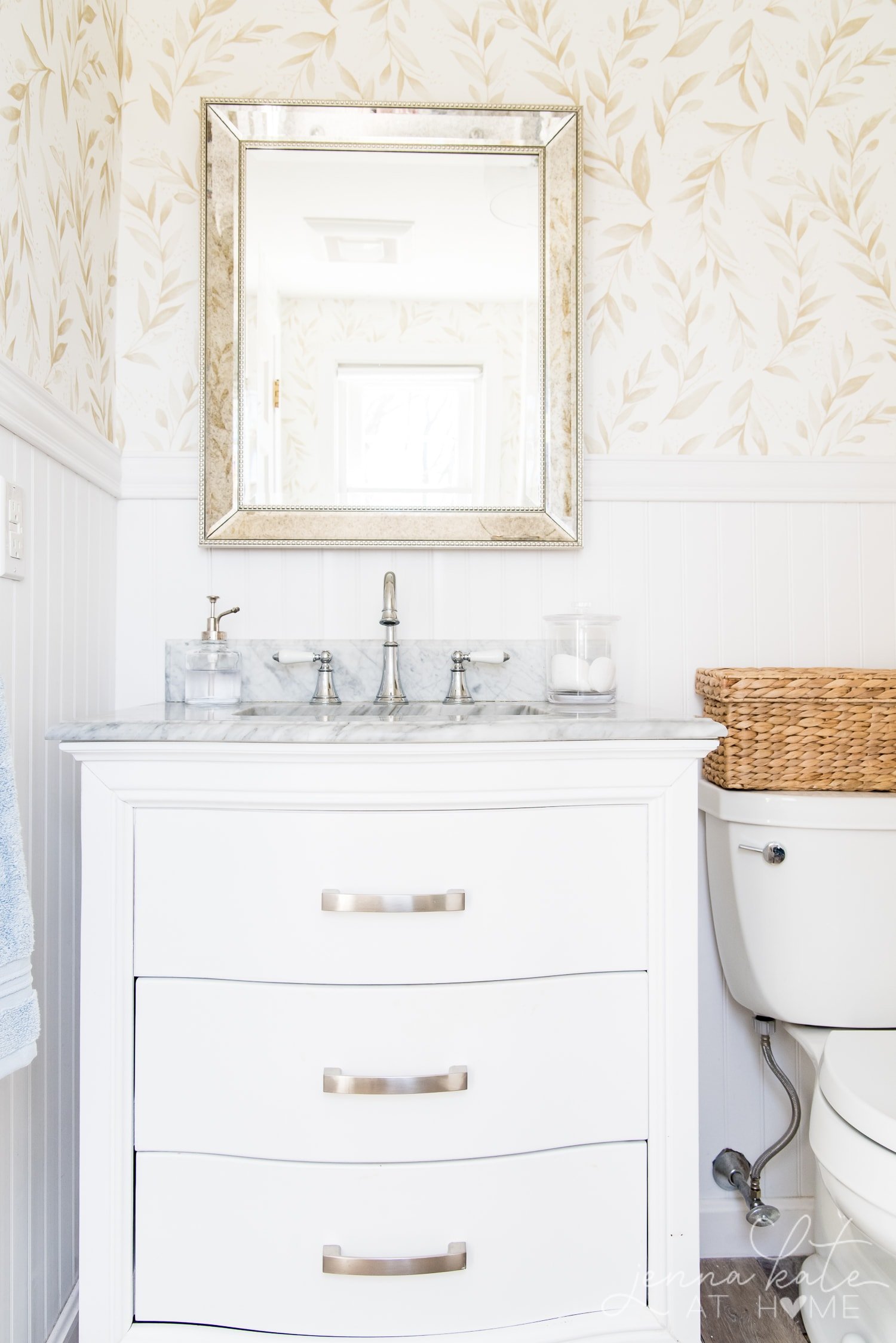 White vanity and marble top with a mercury glass mirror overhead showcasing the elegant floral bathroom wallpaper