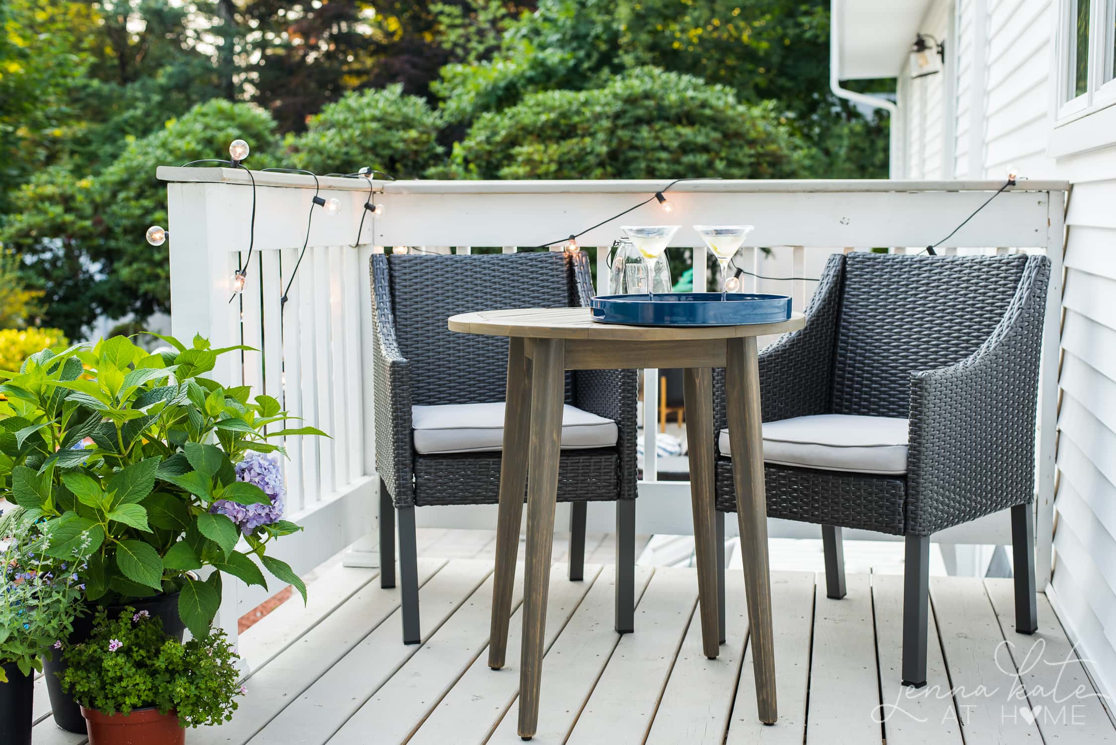 An outdoor deck featuring a pair of grey, wicker chairs with grey seat cushions, and a small table with a blue tray holding two martini glasses