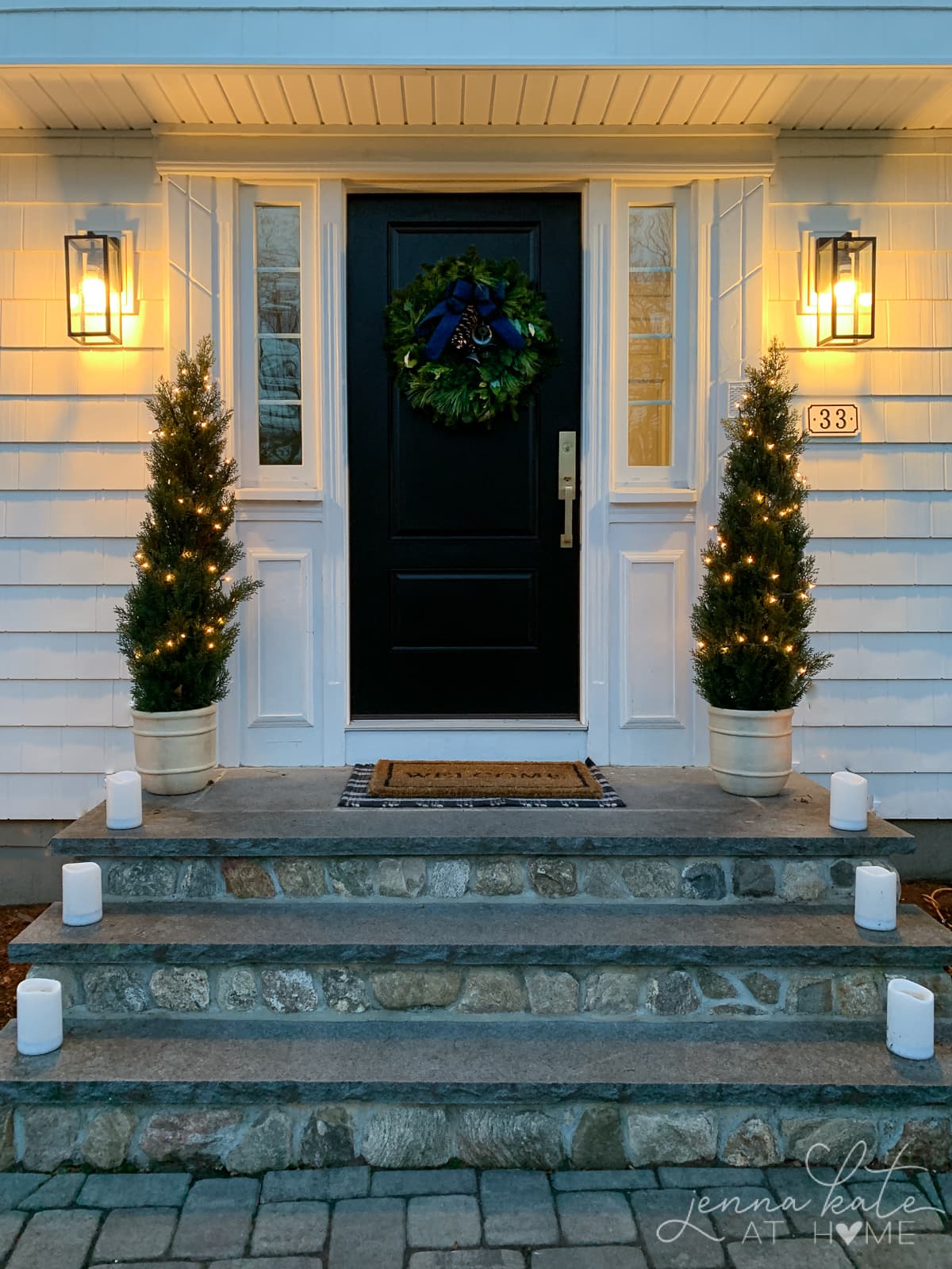 Black front door with a Christmas wreath and two mini trees on the front steps