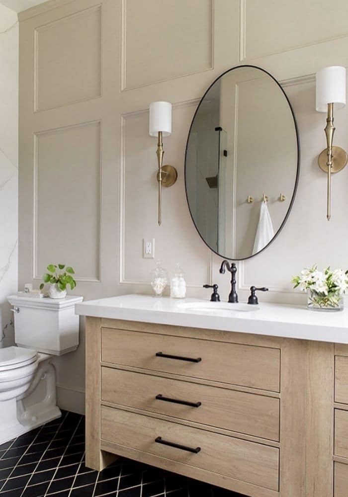 Light wood vanity in a bathroom with wainscoting on the wall that's painted accessible beige, with a black oval mirror
