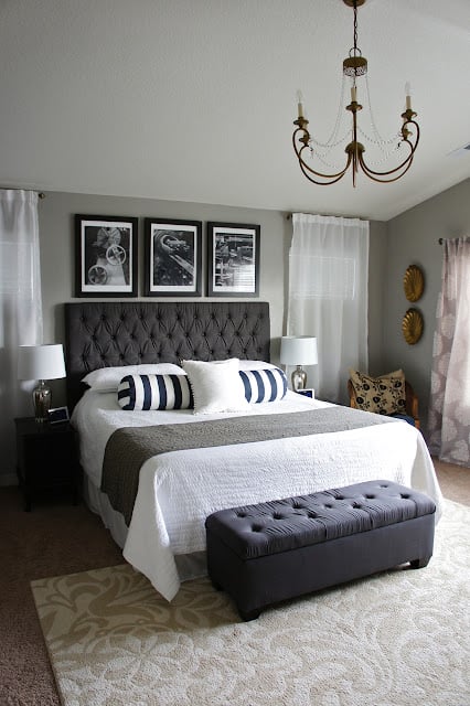 A bedroom with gray walls, an antique chandelier, beige patterned area rug and large bed with dark gray upholstered headboard and grey cushioned bench
