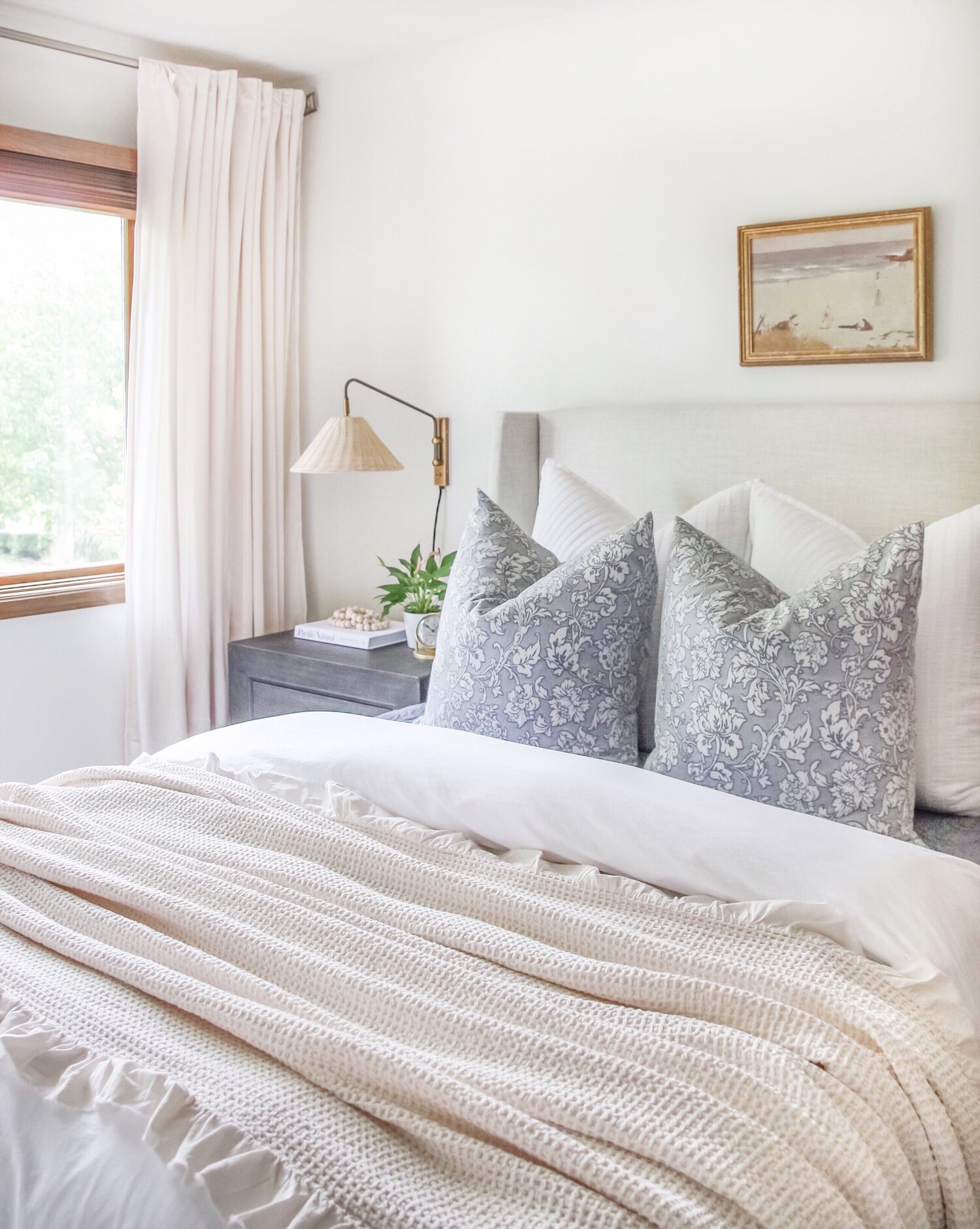 Neutral bedroom with white comforter, beige throw blanket and blue pillows with white curtains and view out the window