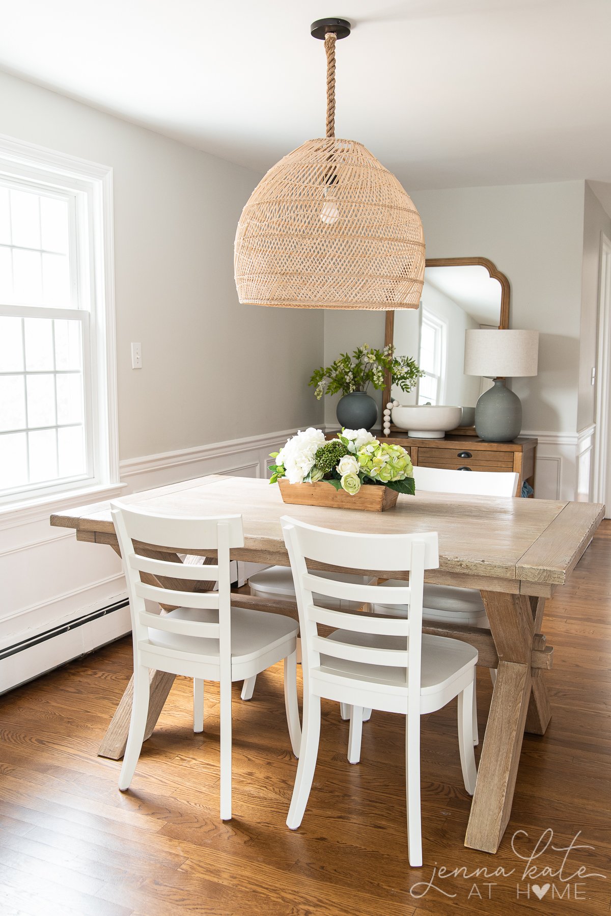 Dining space with wooden table, white chairs and rattan basket pendant. The wainscoting is painted pure white.