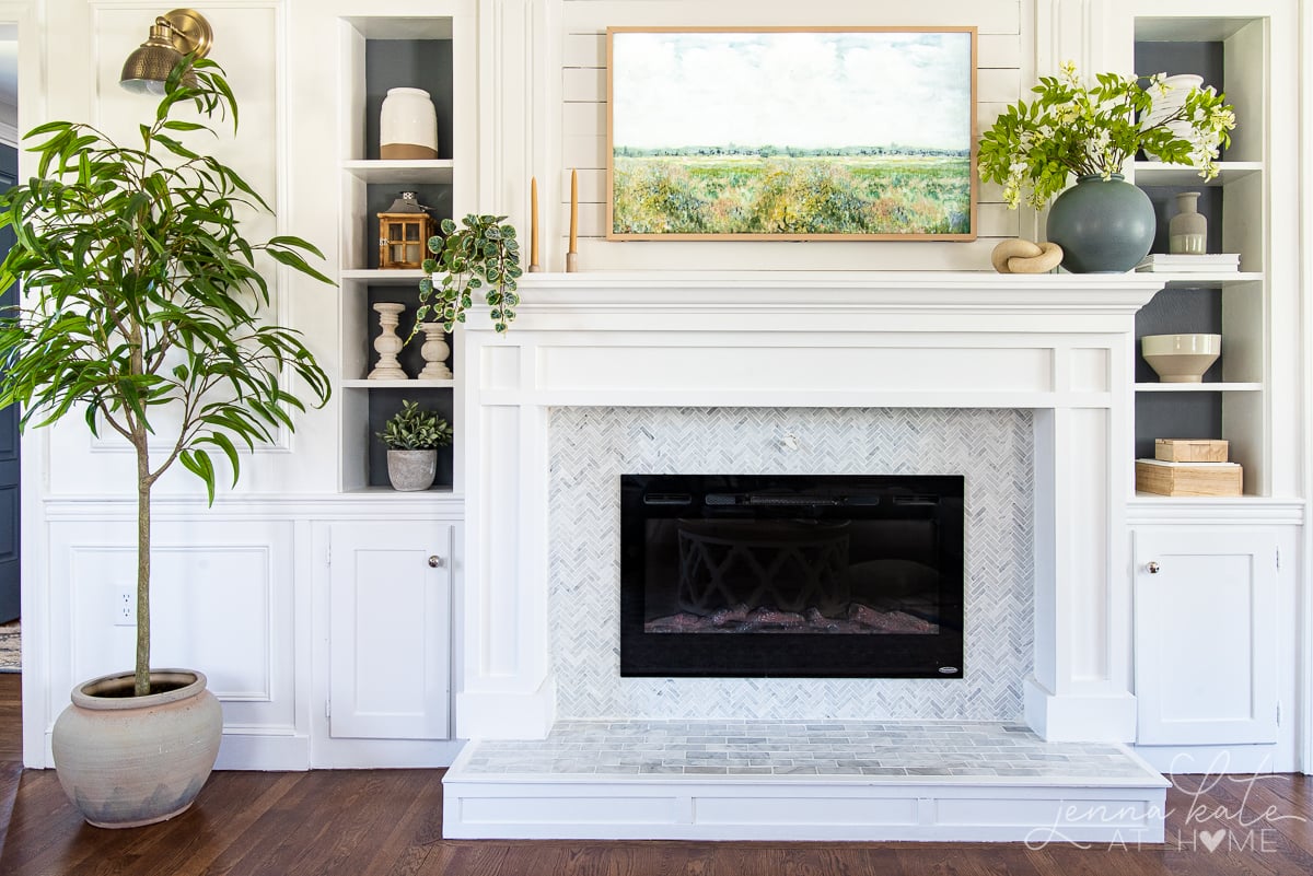 A white painted fireplace with a mantel adorned with faux plants and a frame tv hanging above.
