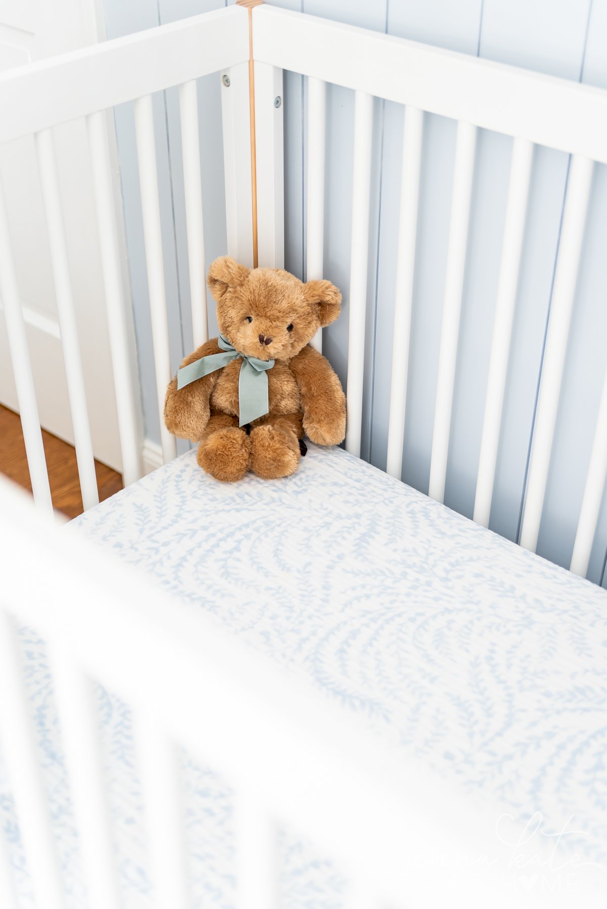 corner of a crib with a teddy bear with a blue bow around his neck