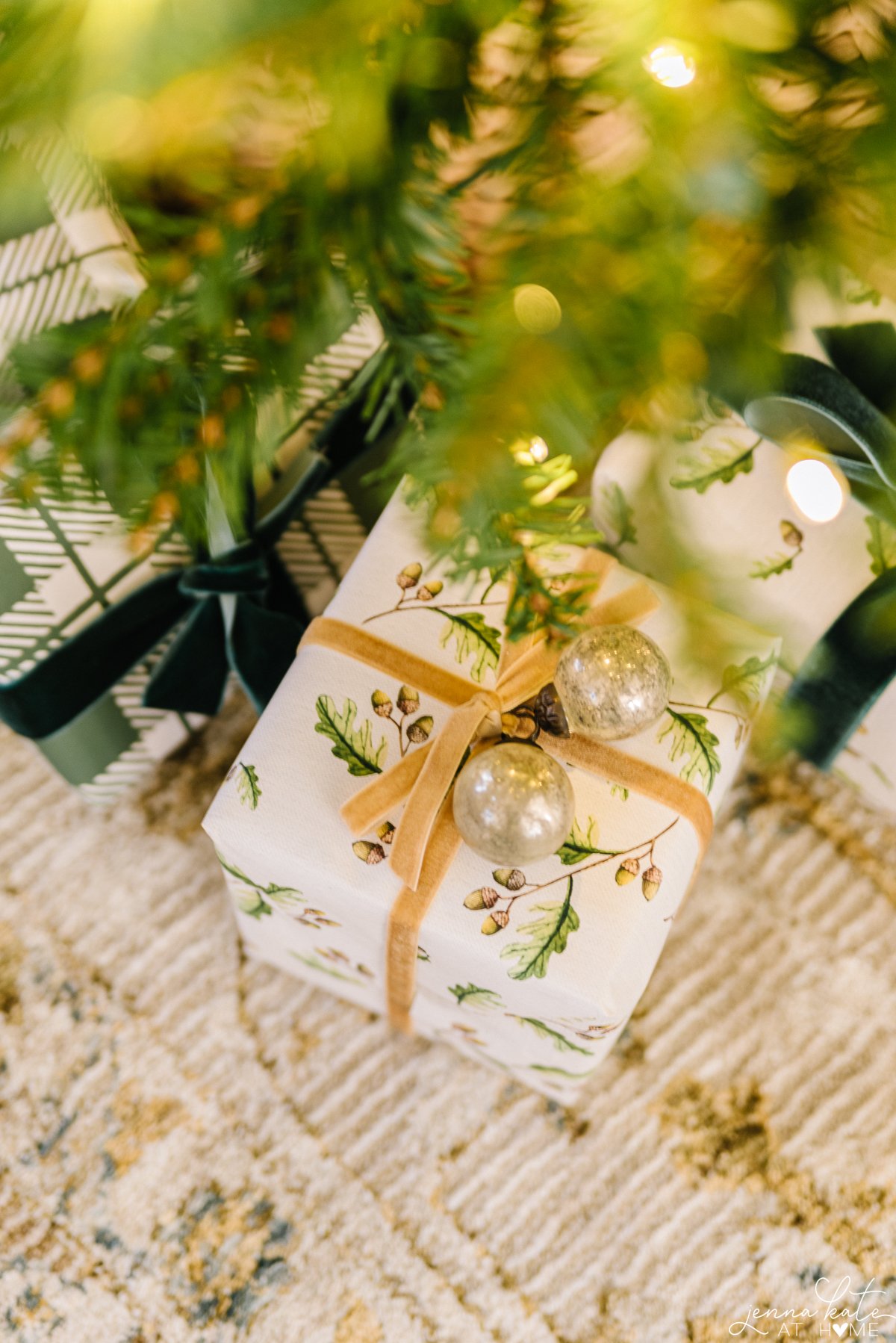 A Christmas gift wrapped in white wrapping paper with an outdoor print of greenery and acorns. A gold ribbon is attached with two small ornaments on the top as decor. The gift is sitting under a Christmas tree.