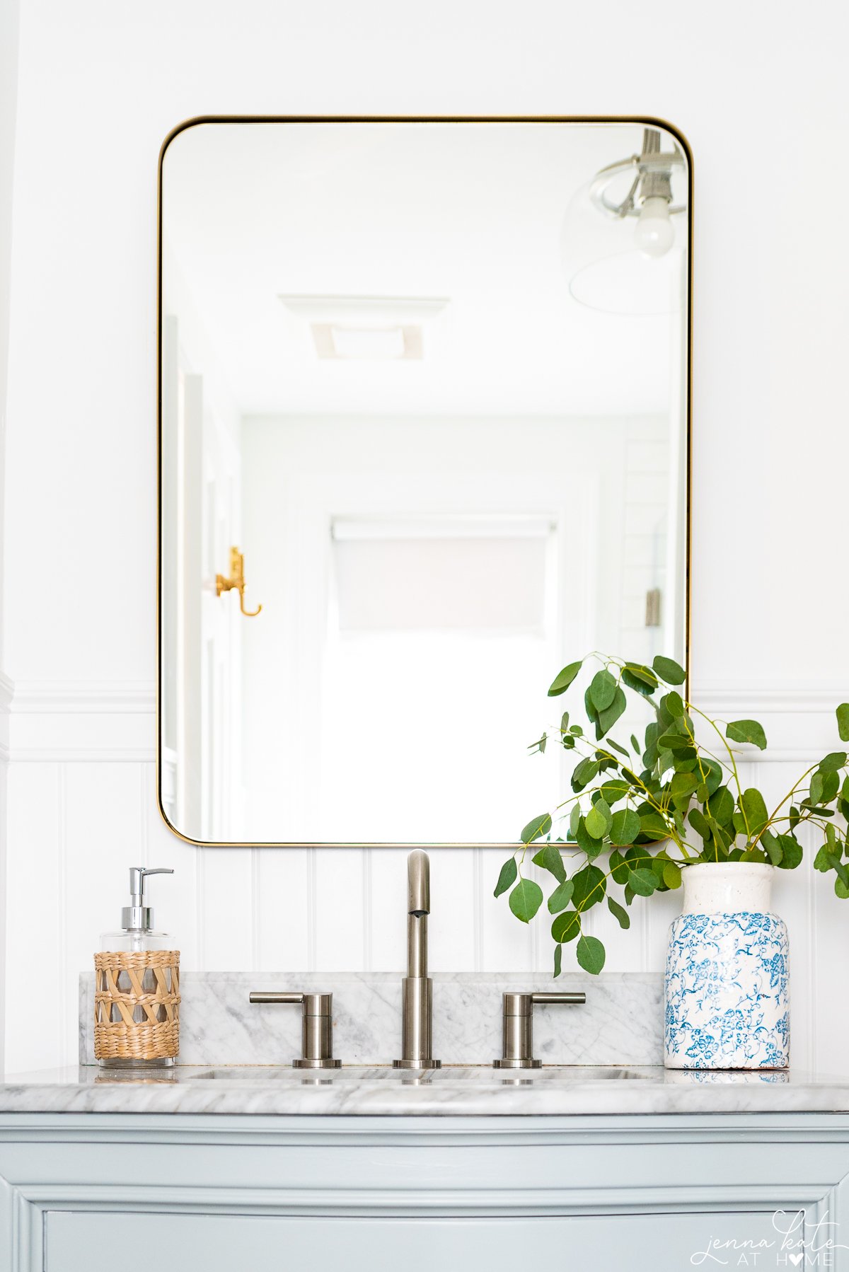 brass mirror over a bathroom vanity with satin nickel faucet and a vase with greenery