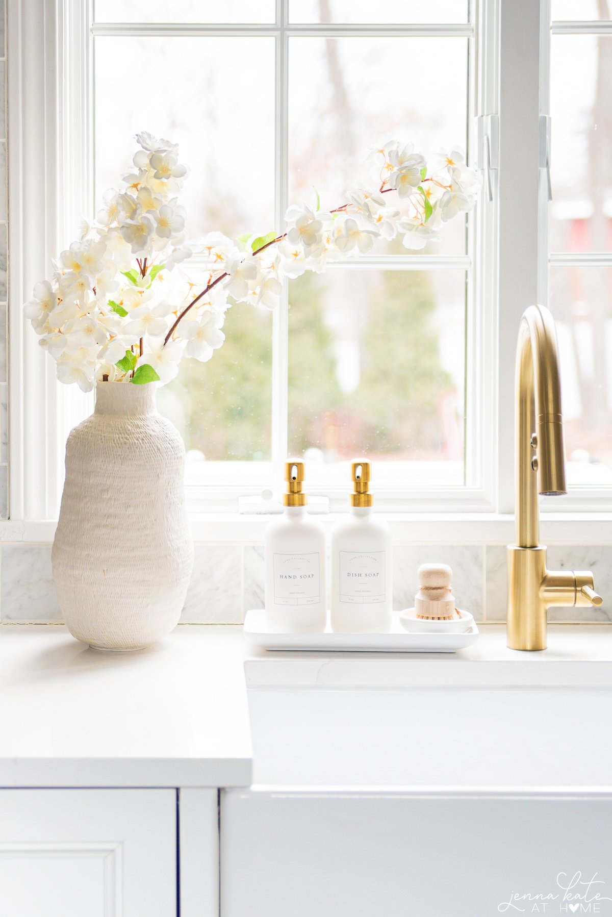 artificial cherry blossoms in a white vase next to a sink with a brass faucet
