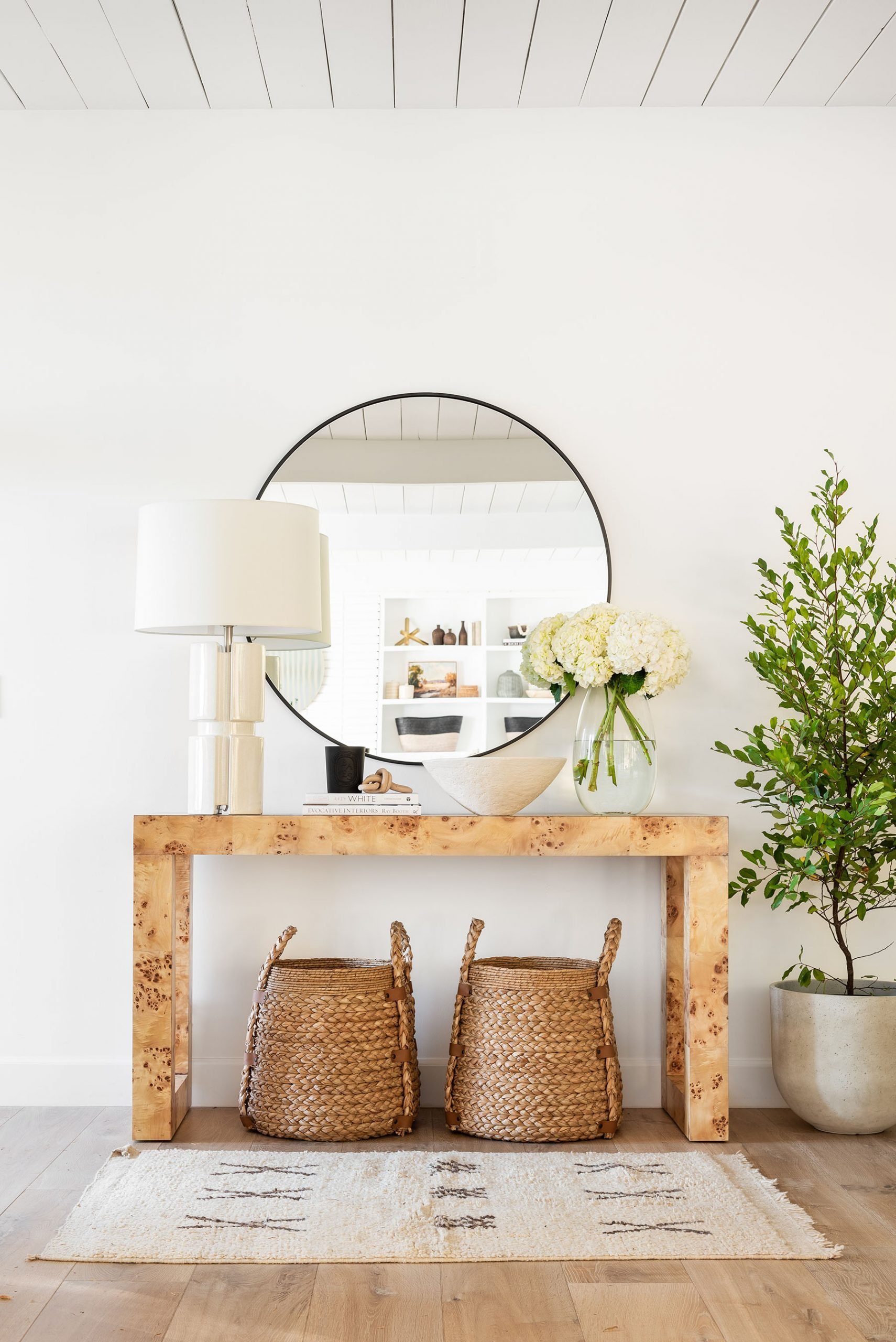 burlwood console table with round mirror, lamp and vase of flowers