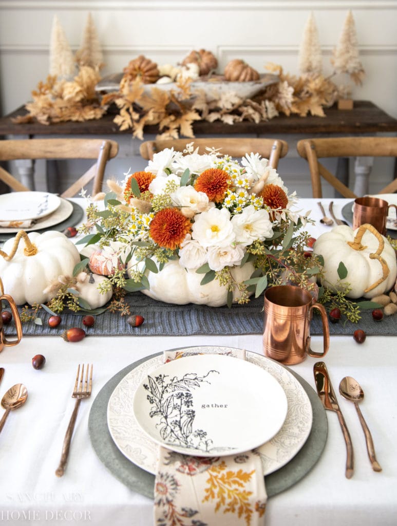 Traditional colors for Thanksgiving on a dining table with pumpkins, floral arrangements, copper cups, and delicate plates. 