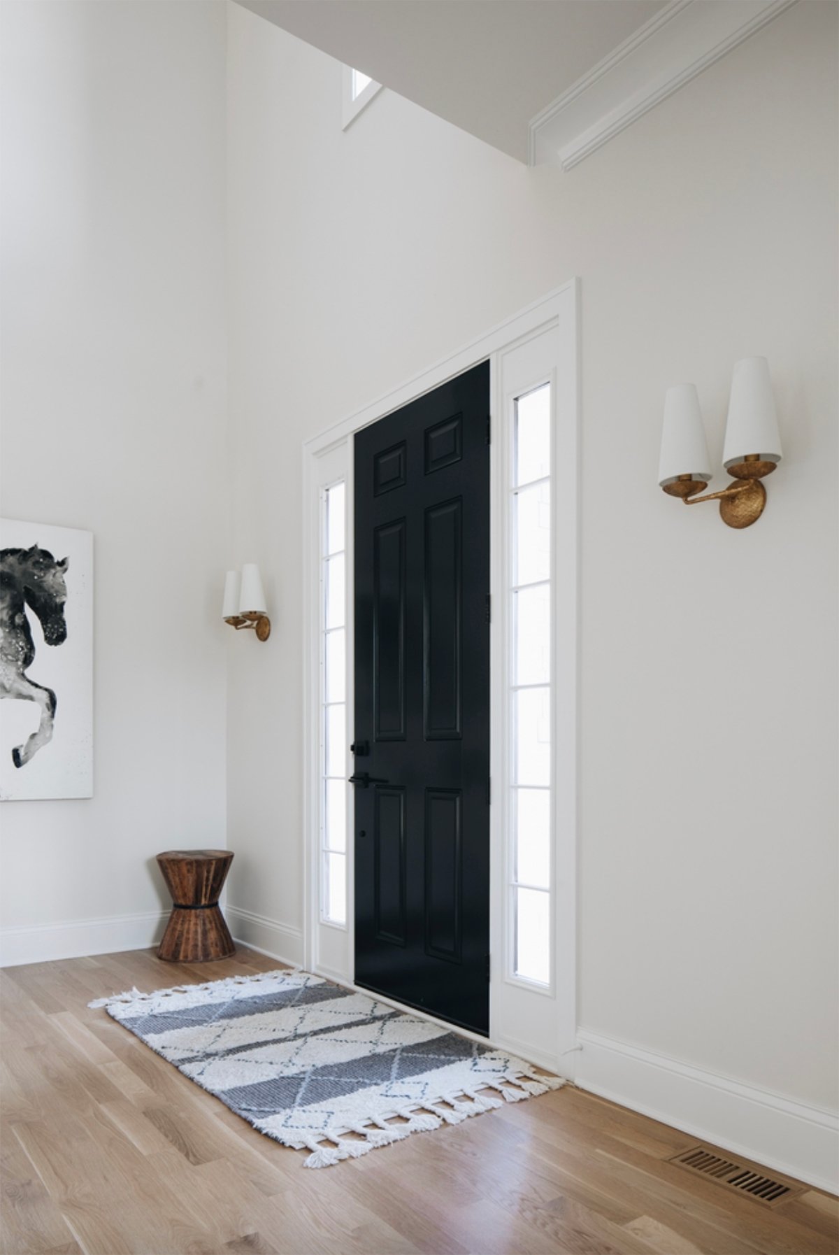 A big entryway with a black door, blue and white mat, and gold light fixtures on the wall. Walls are painted with classic gray.