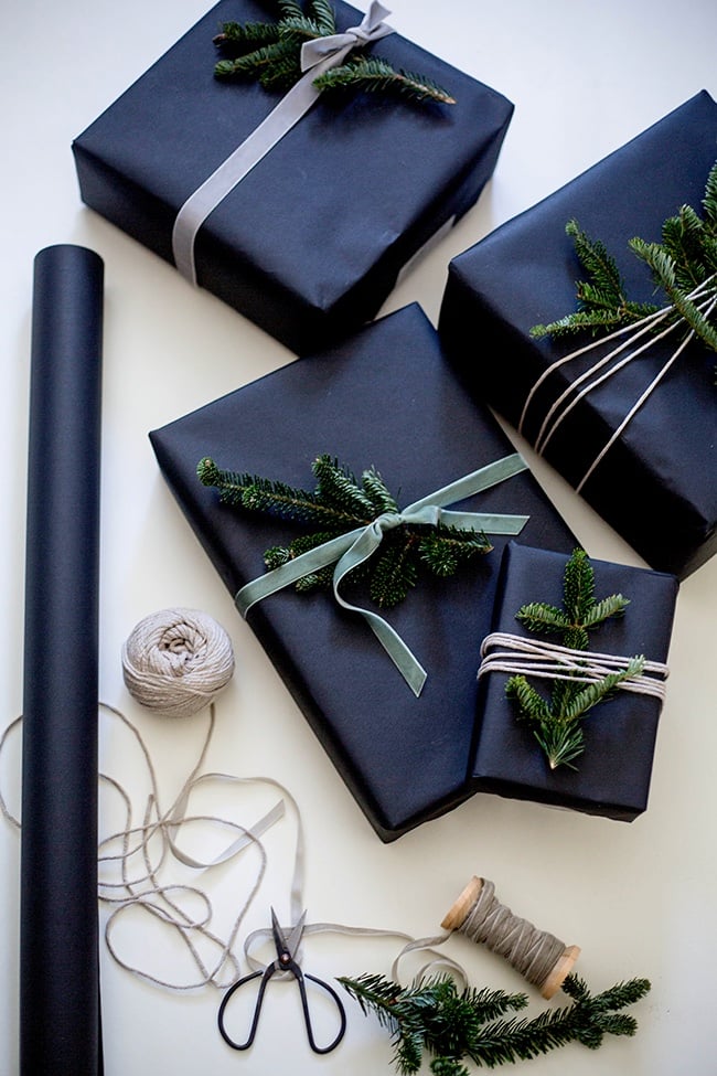 A collection of wrapped presents all in a dark navy paper with greenery on top as decor and sage green ribbon tied around each box.