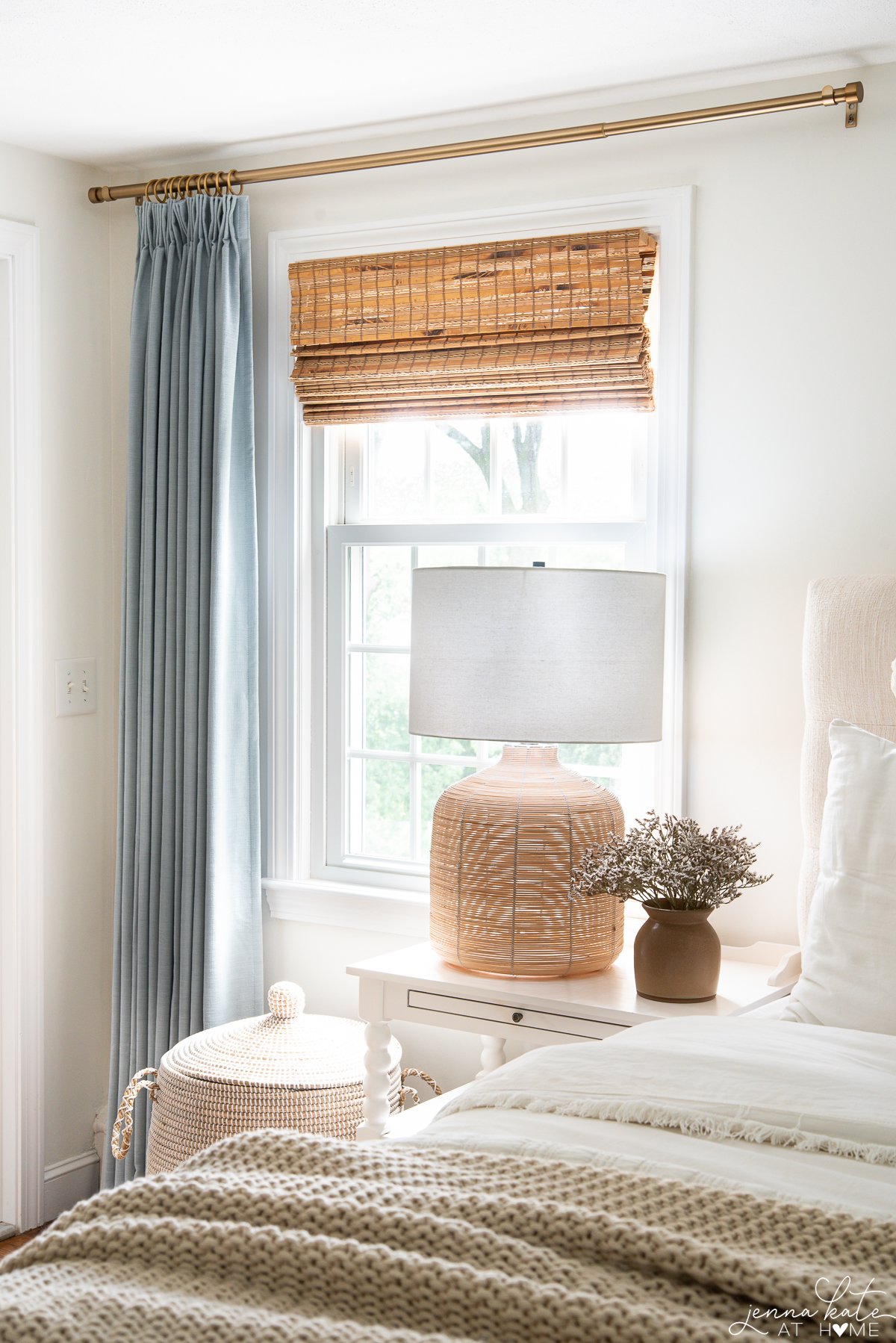 A bedroom window with blue curtains around it and a bedside table in front with a large lamp on display